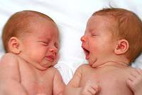 two babies sleeping side by side, one baby yawns while the other baby makes a face